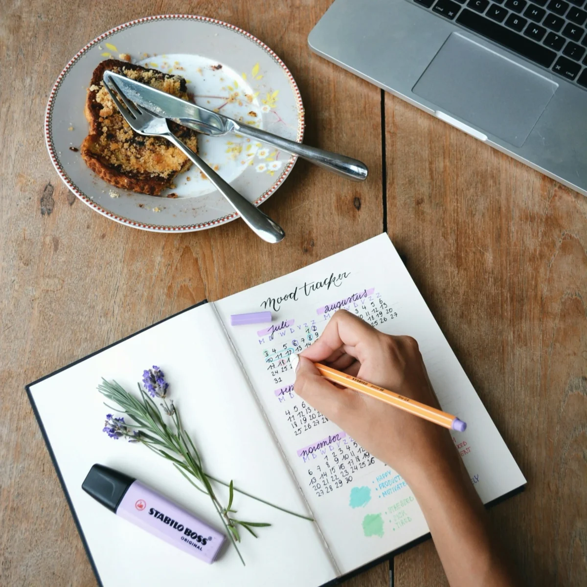 Main qui note ses habitudes dans un carnet posé sur une table en bois, à côté d'une assiette et d'une branche de lavande